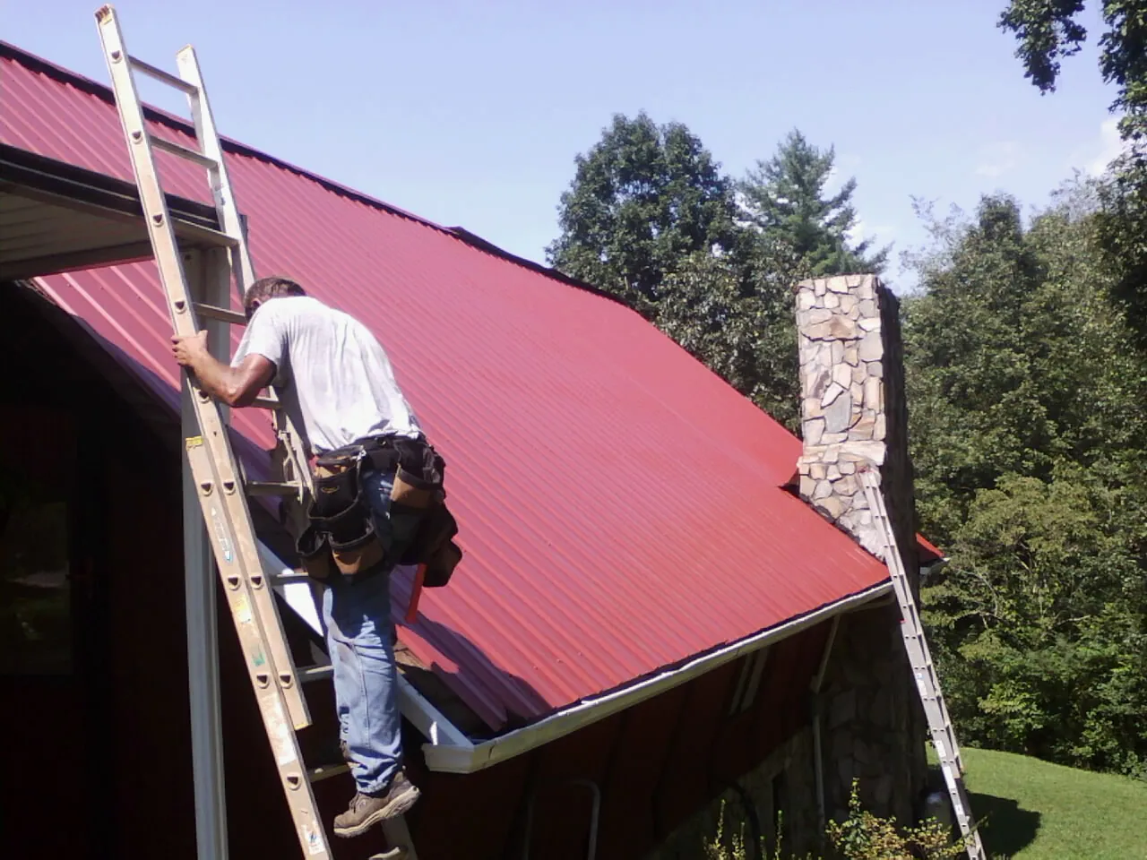 Roofer on a ladder beside a red metal roof and stone chimney — Soffit & Fascia Repair by Roofing Brothers in James Creek